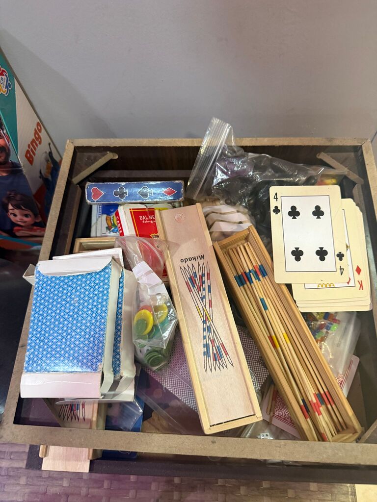 An open wooden box of games: playing cards, pick-up sticks and boxed board games.